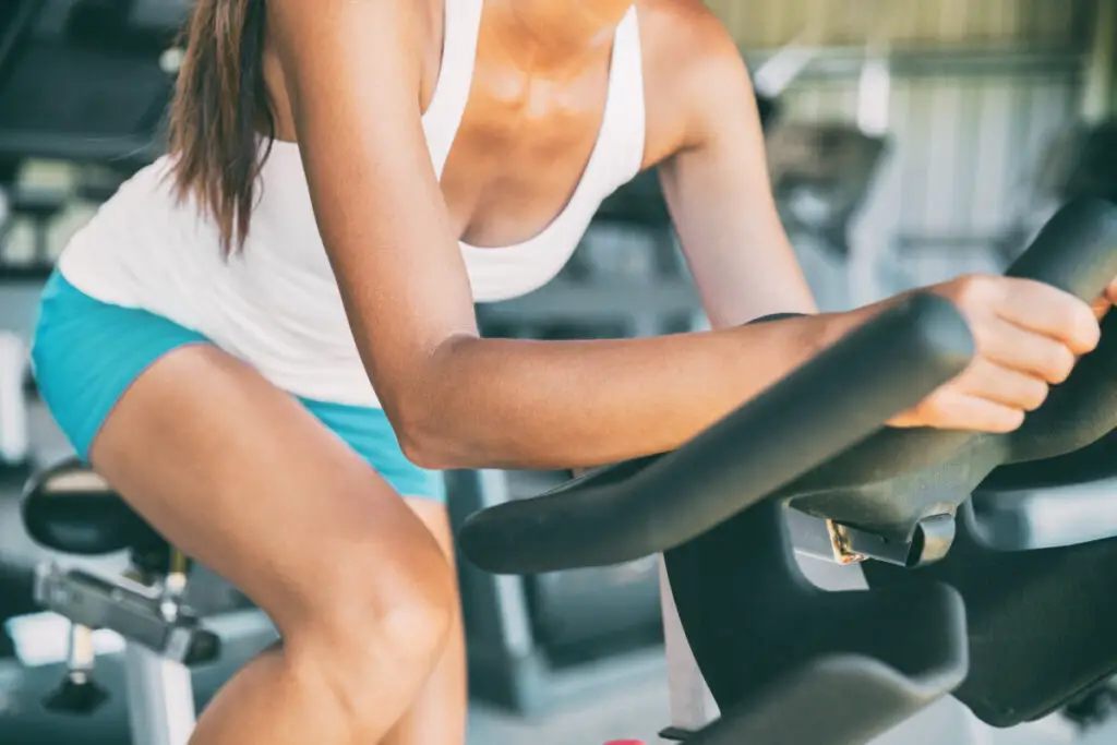 Indoor cycling woman doing HIIT cardio workout biking on indoors gym bike. Girl cyclist working out interval training on bicycle. Closeup of legs and thighs for fat weight loss.